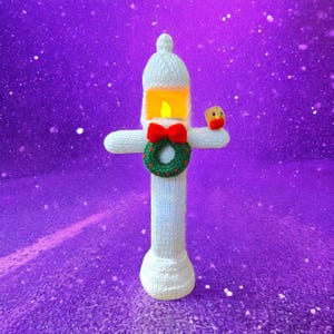 May include: A white knitted Christmas candle decoration, featuring a red bow, a green wreath, and a small bird ornament. The candle has a yellow light inside. The background is purple with white specks.