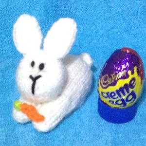 Puede incluir: Un conejito de punto blanco sosteniendo una zanahoria, junto a un huevo de chocolate Cadbury Creme Egg morado, amarillo y azul.