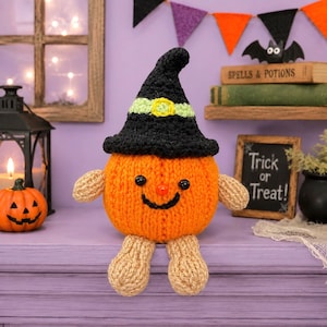 May include: A round, orange knitted pumpkin figure with a black witch hat featuring a light green band and yellow buckle. The figure has tan knitted arms and legs, a small orange nose, and black bead eyes with a stitched black smile.