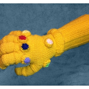 May include: A yellow knitted glove with a detailed hand design and colorful gems on the fingers. The glove is a representation of the Infinity Gauntlet from the Marvel Cinematic Universe.