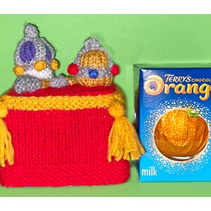 Könnte beinhalten: Ein gestricktes orangefarbenes und graues Spielzeug mit einer roten gestrickten Basis und gelben Quasten. Das Spielzeug steht vor einer Schachtel Terry's Chocolate Orange Milchschokolade.