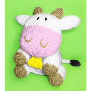 Op de afbeelding: Een wit gebreid knuffeldier in de vorm van een koe met roze oren, een roze gezicht en een gele bel. De koe heeft bruine hoeven en een zwarte staart.