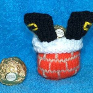 May include: Knitted Christmas stocking with a red brick chimney and a pair of black legs sticking out of the top. The stocking is filled with two gold chocolate balls.