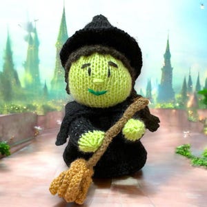 May include: A green knitted witch doll with a black hat and a brown broom. The doll is holding the broom in its right hand and has a friendly expression on its face.