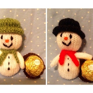May include: Two knitted snowman decorations with green and black hats, red and green scarves, and white faces. Each snowman is holding a gold chocolate ball.