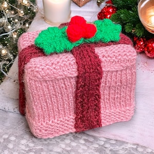 Op de afbeelding: Een roze gebreide cadeaudoos met een rood lint en een groene hulsttak met een rode bes erop.