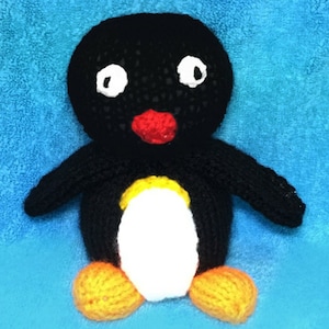 Puede incluir: Un juguete de pingüino de punto negro y blanco con un pico rojo y patas amarillas. El pingüino está sentado en una toalla azul.