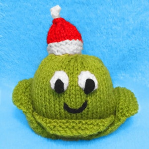 May include: A green knitted Christmas ornament shaped like a creature with a red and white Santa hat. The ornament has white eyes, black pupils, and a black smiling mouth.