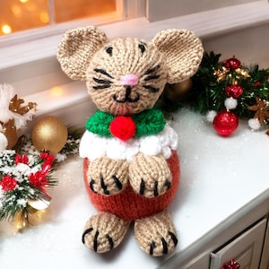 May include: A knitted mouse wearing a green and red Christmas-themed hat and scarf. The mouse is sitting on a brown and white knitted base that resembles a Christmas pudding.