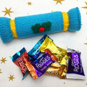 Puede incluir: Un cracker navideño de punto azul con ribete amarillo y diseño de acebo. El cracker está lleno de varios caramelos de chocolate envueltos individualmente, cada uno con la palabra "Roses" impresa en el envoltorio.