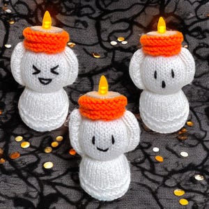 May include: Three knitted ghost figures in white, each with an orange candle hat and a smiling or surprised face. They have small, flickering LED lights on top, mimicking candle flames. The background is dark fabric with gold confetti.