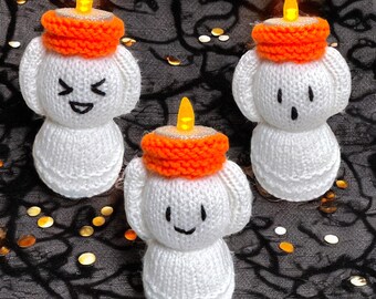 May include: Three handmade, white knitted ghost figures with orange candle hats and smiling or surprised faces. Each ghost has a small, flickering LED light at the top, resembling a candle flame. The background is a dark fabric with gold confetti.