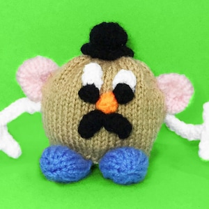 Puede incluir: Un juguete de patata de punto con un sombrero de copa negro, cuerpo marrón, orejas rosas, brazos blancos y zapatos azules. La patata tiene una cara blanca con ojos negros, un bigote negro y una nariz naranja.