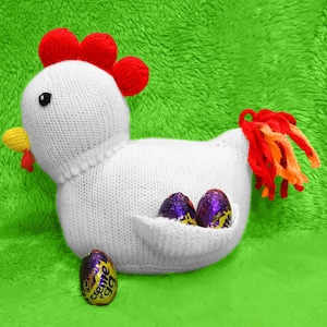 May include: White knitted chicken with a red comb and tail feathers holding three purple and gold chocolate eggs. The chicken is sitting on a green fuzzy surface.