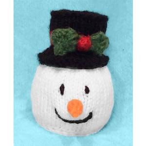May include: A white knitted snowman ornament with a black top hat, green and red bow, and a black stitched smile. The snowman has a black stitched nose and orange felt nose.