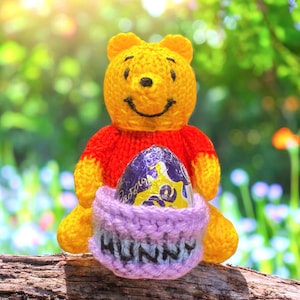 Könnte beinhalten: Ein handgefertigtes gelb-rotes Winnie-the-Pooh-Plüschtier, das ein lila Osterei hält. Das Plüschtier sitzt auf einer Holzoberfläche, mit einem gestrickten Korb mit der Aufschrift "HUNNY". Der Hintergrund ist verschwommen grün und gelb.