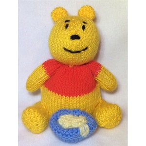 Puede incluir: Un oso de peluche amarillo tejido que lleva una camisa roja y amarilla, sentado con un tazón azul de miel delante de él.