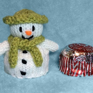 May include: A white knitted snowman with a green hat and scarf, holding a red and silver wrapped chocolate.