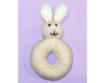 May include: A knitted cream-coloured bunny rattle with light blue ears and a pink nose. The rattle is shaped like a ring.