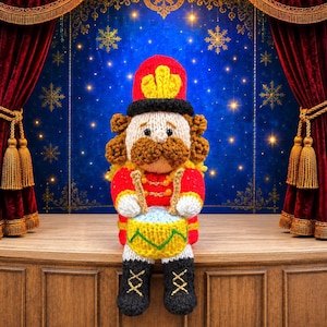Könnte beinhalten: Eine gehäkelte Nussknacker-Puppe in einer roten Uniform mit goldenen Akzenten, einem hohen roten Hut und schwarzen Stiefeln. Die Puppe hat ein beiges Gesicht, braunes Haar, einen braunen Schnurrbart und hält zwei Trommelstöcke über einer gelben Trommel.