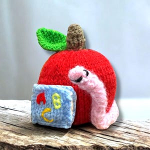 May include: A red knitted apple with a green leaf and a pink knitted worm peeking out. The apple is holding a blue book with the letters A, B, and C embroidered on it.