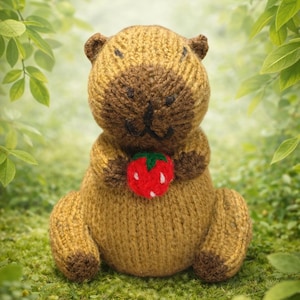 May include: A knitted bear in brown and tan tones, clutching a red strawberry with green leaves and white details. The bear features a black stitched nose and eyes. The backdrop is a soft green with foliage.