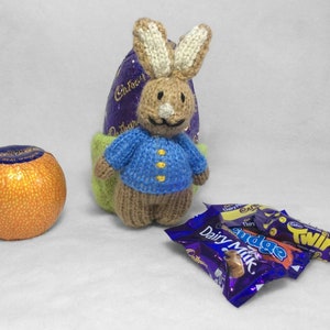 Puede incluir: Un conejito de punto con un jersey azul se sienta dentro de un huevo de Pascua verde y morado. El conejito sostiene un huevo de chocolate morado. Hay tres barras de chocolate en la mesa: Cadbury Dairy Milk, Cadbury Twirl y Cadbury Fudge.