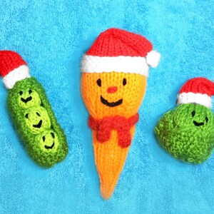 May include: Three knitted Christmas ornaments: a carrot with a red Santa hat, a green pea pod with three peas, and a green ball with a Santa hat. All three ornaments have smiling faces.