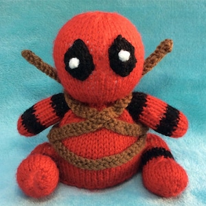 May include: A red and black knitted plush toy of Deadpool, a superhero character, is tied up with brown yarn. The toy has a black eye mask with white eyes.