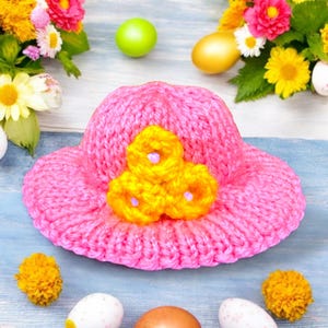 May include: A pink knitted hat with three yellow flower embellishments. The hat is on a blue wooden surface with Easter eggs and flowers.