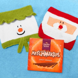 May include: A green and white knitted snowman ornament and a red and white knitted Santa Claus ornament, both with embroidered faces. A box of Quality Street Matchmakers Zingy Orange sweets is in the foreground.