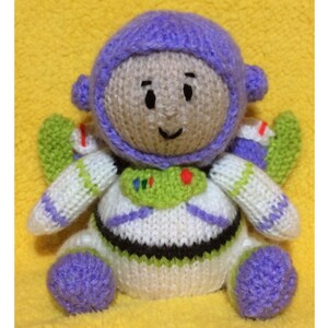 Puede incluir: Un juguete de punto de Buzz Lightyear de la franquicia Toy Story. El juguete es morado, blanco y verde. Tiene un sombrero morado y un chaleco verde con un corazón rojo.