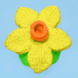 May include: A knitted daffodil flower with a yellow body and an orange centre. The flower is sitting on a green stem.