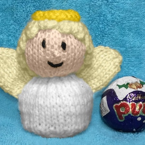 May include: A white knitted angel with a yellow halo and a pink face. The angel is holding a purple chocolate egg with the word "Puds" on it.