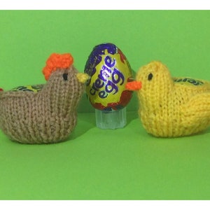 Puede incluir: Dos decoraciones de Pascua de punto, una gallina marrón y un pato amarillo, junto a un huevo de chocolate Creme Egg morado y dorado.