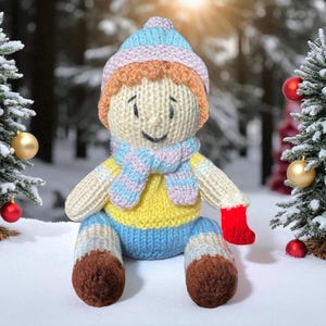 May include: A handmade knitted doll wearing a striped blue and pink hat, scarf, and blue pants. The doll has a yellow sweater, red mitten, and brown boots. The doll is sitting in the snow near decorated Christmas trees.