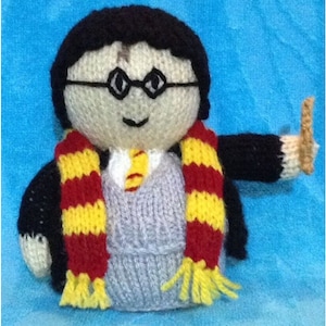May include: Knitted Harry Potter doll wearing a black robe, red and yellow striped scarf, and round glasses. The doll is holding a brown wand.