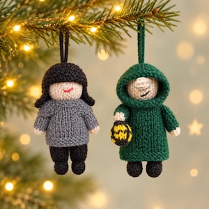 May include: Two handmade crochet ornaments hanging from a branch. One figure has black hair, a gray sweater, and black pants. The second figure wears a dark green hooded cloak and holds a small yellow and black patterned lantern in one hand.