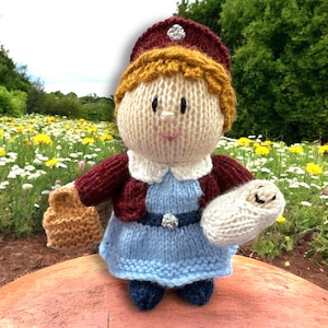 May include: A knitted doll wearing a blue dress, a maroon cardigan, and a red hat with a silver button. The doll is holding a knitted baby doll in one arm and a brown knitted bag in the other.