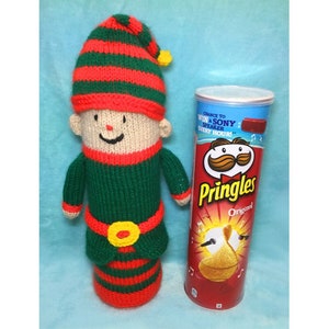 May include: A knitted elf doll wearing a green and red striped hat and a green and red striped outfit. The elf is standing next to a can of Pringles potato chips. The can has a red background with the Pringles logo and the text "Original".