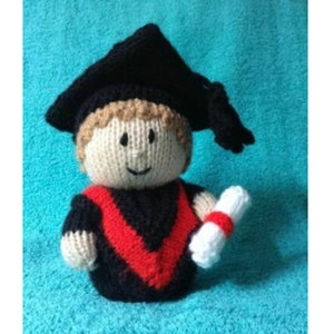 May include: A knitted graduation doll wearing a black cap and gown with a red sash and holding a white scroll.