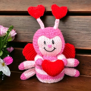 May include: A pink and red knitted bee with heart-shaped wings and antennas holding a red heart. The bee has a smiling face and is sitting on a wooden surface.