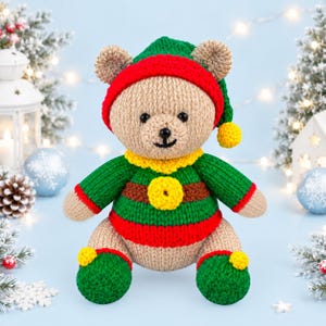 Può includere: Un orsacchiotto all'uncinetto che indossa un maglione a righe verdi e rosse con colletto e bottone centrale gialli, un cappello da elfo verde e rosso con un pon pon giallo e scarpe verdi con pon pon gialli. L'orso ha una pelliccia in filato marrone chiaro.