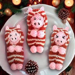 May include: Three knitted pig ornaments in a sleeping pose. Each pig is wearing a red and white striped sweater and has a pink snout and hooves.