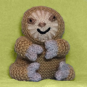 May include: A knitted sloth toy with brown fur, a light brown face, and purple claws. The sloth is sitting with its arms crossed and has a happy expression.