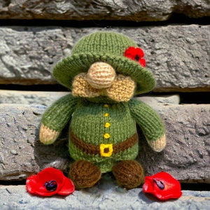 May include: A knitted toy soldier wearing a green hat with a red poppy, a green uniform with brown boots, and a brown belt with a yellow buckle. The toy is standing on a stone surface with two red poppies on either side.