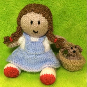 May include: A knitted doll with brown hair, a blue and white dress, and red shoes. The doll is holding a brown knitted basket with a brown knitted dog inside.