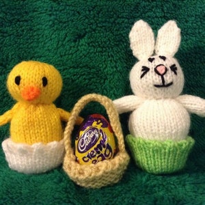 May include: A knitted yellow chick, a knitted white bunny, and a small knitted basket with a Cadbury Creme Egg inside. The toys are sitting on a green fuzzy surface.