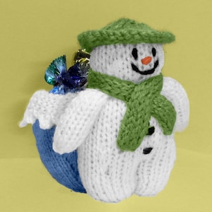 Op de afbeelding: Een gebreide sneeuwpop met een groene hoed en sjaal, die een blauw en groen ingepakt cadeau vasthoudt. De sneeuwpop is wit met zwarte ogen en een rode neus.