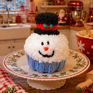 Peut inclure: Une décoration de cupcake bonhomme de neige crochetée avec une tête blanche duveteuse, un haut-de-forme noir avec une bande rouge et des feuilles de houx vertes, un nez orange et un sourire noir. La base est une texture côtelée bleu clair.